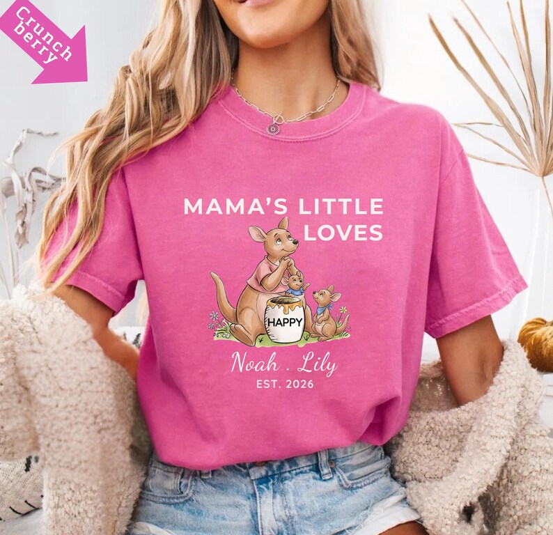 Puede incluir: Una camiseta rosa con el texto "MAMA'S LITTLE LOVES" y un dise&ntilde;o de familia de canguros de dibujos animados. El dise&ntilde;o incluye un canguro, dos cr&iacute;as y un tarro de miel con la palabra "HAPPY". Los nombres "Noah. Lily" y el a&ntilde;o "EST. 2026" tambi&eacute;n est&aacute;n impresos en la camiseta.