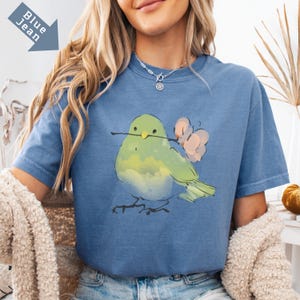May include: Blue jean colored t-shirt featuring a watercolor illustration of a green bird holding a twig and flowers. The bird is light green and yellow with a light pink flower. The shirt has the words "Blue Jean" printed on it.