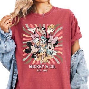 Puede incluir: Camiseta roja con un gr&aacute;fico de Mickey Mouse, Minnie Mouse, Donald Duck, Daisy Duck, Goofy y Pluto. El dise&ntilde;o est&aacute; sobre un fondo de rayas retro. El texto "MICKEY & CO. EST. 1928" est&aacute; impreso debajo de los personajes.