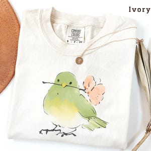 May include: Ivory-colored t-shirt with a watercolor illustration of a green bird holding a twig and a pink flower. The shirt has a crew neck and short sleeves. A gold necklace with a pendant is draped on the shirt.