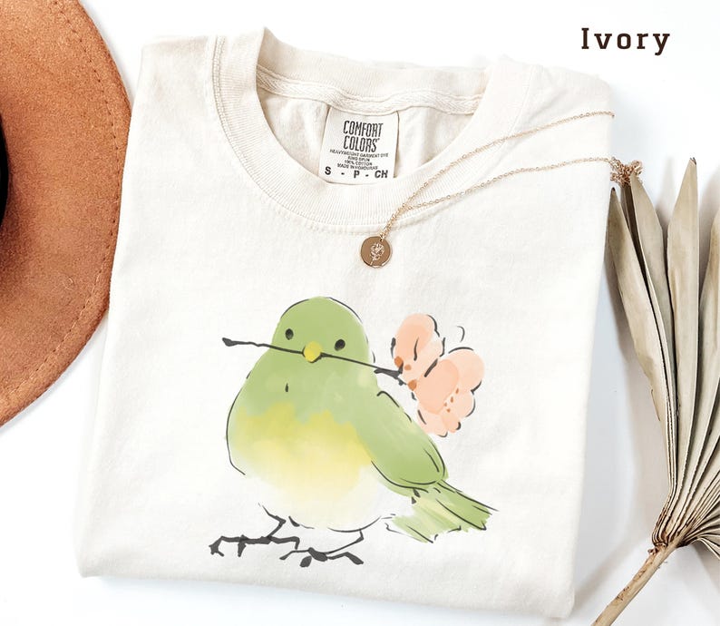 May include: Ivory-colored t-shirt with a watercolor illustration of a green bird holding a twig and a pink flower. The shirt has a crew neck and short sleeves. A gold necklace with a pendant is draped on the shirt.