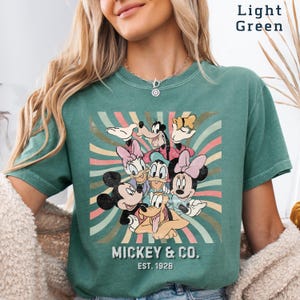 Puede incluir: Camiseta verde claro con un dise&ntilde;o retro de Mickey Mouse y sus amigos. El dise&ntilde;o incluye a Mickey, Minnie, Donald, Daisy, Goofy y Pluto, con el texto "Mickey & Co. Est. 1928". El fondo tiene rayas coloridas.