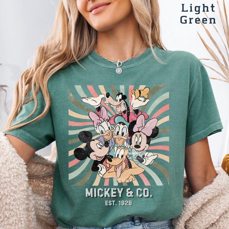 Puede incluir: Camiseta verde claro con un dise&ntilde;o retro de Mickey Mouse y sus amigos. El dise&ntilde;o incluye a Mickey, Minnie, Donald, Daisy, Goofy y Pluto, con el texto "Mickey & Co. Est. 1928". El fondo tiene rayas coloridas.