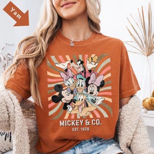 Puede incluir: Camiseta naranja de manga corta con un gr&aacute;fico de Mickey Mouse y sus amigos. El dise&ntilde;o incluye a Mickey, Minnie, Donald, Daisy, Goofy y Pluto, con el texto "Mickey & Co. Est. 1928". El fondo tiene rayas retro.