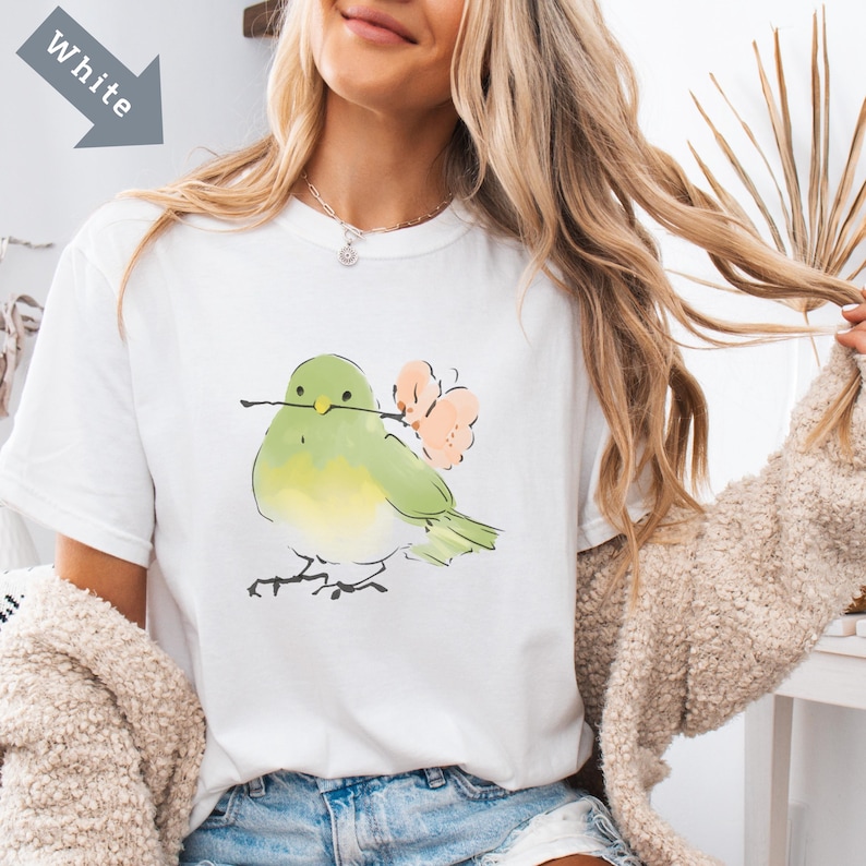 May include: White t-shirt featuring a watercolor illustration of a green and yellow bird holding a twig and flowers. The bird is perched on thin black lines. The word "White" is in a gray arrow in the upper left corner.