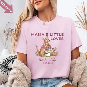 Puede incluir: Camiseta rosa claro con el texto "MAMA'S LITTLE LOVES" y un gr&aacute;fico de familia de canguros. El gr&aacute;fico incluye un canguro, dos cr&iacute;as y un tarro de miel. Debajo del gr&aacute;fico est&aacute;n los nombres "Noah . Lily" y el a&ntilde;o "EST. 2026".