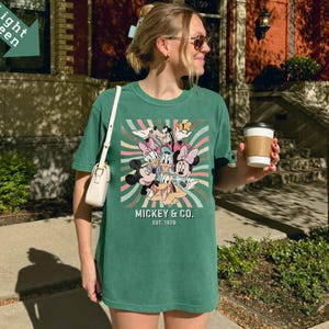 Puede incluir: Una camiseta verde con un estampado de Mickey Mouse y sus amigos, incluyendo a Mickey, Minnie, Donald, Daisy y Goofy. El texto "Mickey & Co. Est. 1928" tambi&eacute;n es visible. La camiseta tiene un corte relajado.