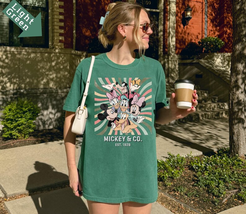 Puede incluir: Una camiseta verde con un estampado de Mickey Mouse y sus amigos, incluyendo a Mickey, Minnie, Donald, Daisy y Goofy. El texto "Mickey & Co. Est. 1928" tambi&eacute;n es visible. La camiseta tiene un corte relajado.