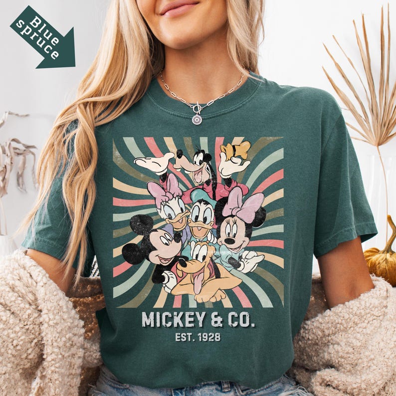 Puede incluir: Camiseta de color verde azulado con un dise&ntilde;o retro de Mickey Mouse y sus amigos. El gr&aacute;fico incluye a Mickey, Minnie, Donald, Daisy, Goofy y Pluto, con el texto "Mickey & Co. Est. 1928". El dise&ntilde;o tiene un aspecto vintage y desgastado con rayas coloridas.