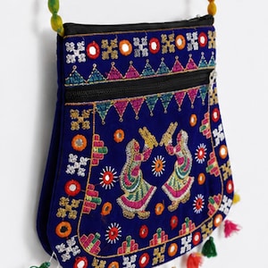 May include: A royal blue crossbody bag featuring intricate multi-colored embroidery, small circular mirrors, and two figures dancing. The bag has a black zippered closure, a rainbow-colored beaded strap, and several small colorful tassels hanging.