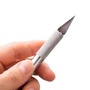 May include: A silver craft knife with a textured handle and a sharp, angled blade. The blade is made of dark metal. The handle is silver with a textured grip. The knife is designed for precision cutting tasks.