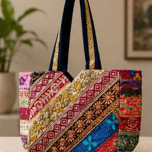 Puede incluir: Un bolso tote con un diseño de patchwork de paneles de tela bordados multicolores. El bolso tiene asas azul oscuro con ribete dorado y muestra intrincados patrones florales, formas geométricas y pequeños espejos circulares en toda la superficie.