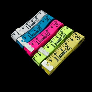 May include: Four colorful Fargo measuring tapes in white, blue, pink, and yellow. Each tape is marked with black numbers and measurement increments. The tapes are arranged diagonally against a black background.