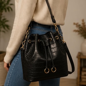 May include: A black bucket bag with a textured crocodile-embossed pattern. The bag features a drawstring closure with gold-toned metal eyelets and rings, a matching black shoulder strap, and gold-toned hardware connecting the strap to the bag.