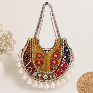 May include: A crescent-shaped shoulder bag featuring a patchwork design of yellow, red, and dark blue fabric with floral embroidery, small circular mirrors, and white cowrie shells. The bag is finished with white fabric tassels along the bottom.