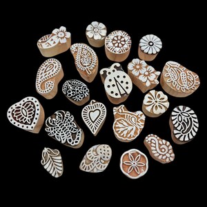 May include: A collection of wooden printing blocks with various designs, including flowers, paisley, and a ladybug. The blocks are light brown with white or black patterns, arranged against a black background. These blocks are likely used for textile printing or crafting.