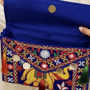 May include: A royal blue clutch bag featuring intricate multi-colored embroidery of an elephant, accented with small circular mirrors, metallic gold-tone coins, and three hanging tassels in orange, dark red, and green, with a magnetic snap closure.