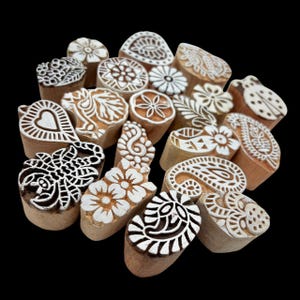 Puede incluir: Una colección de bloques de impresión de madera con varios diseños tallados, incluyendo flores, paisley y patrones geométricos. Los bloques son de color marrón claro con diseños blancos, dispuestos sobre un fondo negro. Las formas varían.