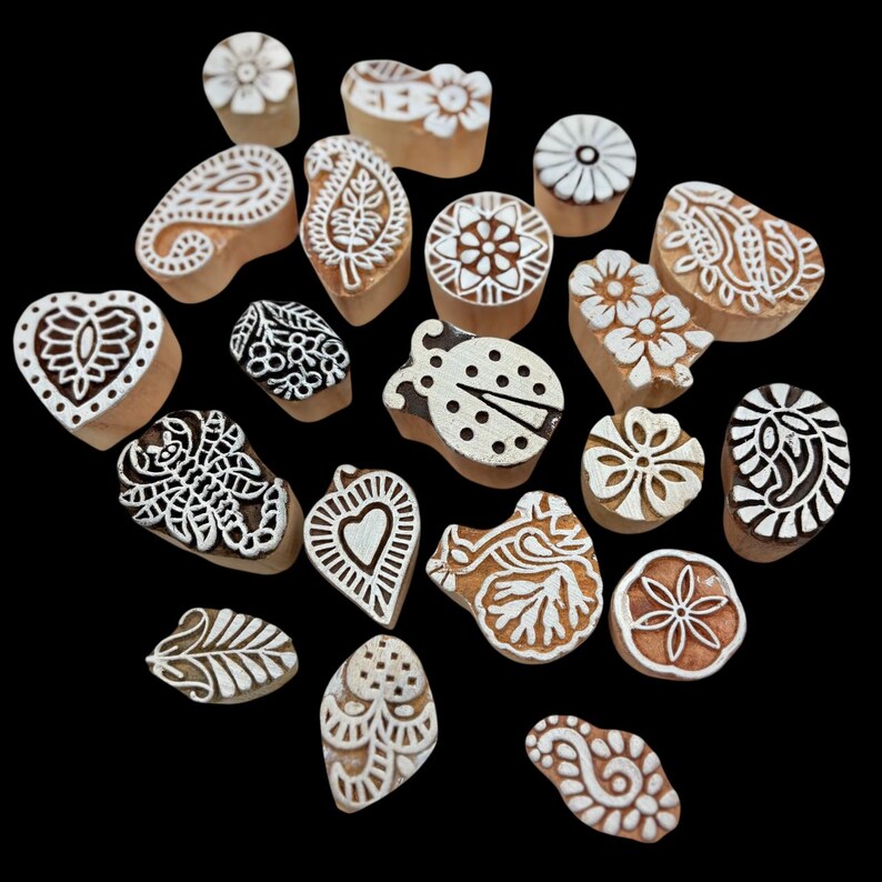 May include: A collection of wooden printing blocks with various carved designs, including flowers, paisley, hearts, and a ladybug. The blocks are light brown with white patterns, set against a black background.