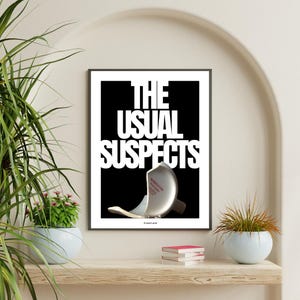 May include: A framed art print with the words "THE USUAL SUSPECTS" in white block letters against a black background. A broken, metallic-coloured object is featured below the text. The print is displayed on a wooden shelf with plants.