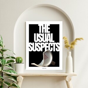 May include: A framed art print with the text "THE USUAL SUSPECTS" in white against a black background. A broken object is featured below the text. The print is displayed on a wooden shelf with plants and a vase.