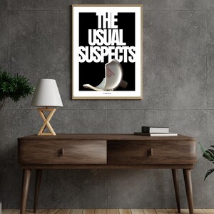 May include: A framed art print featuring the words "THE USUAL SUSPECTS" in white on a black backdrop. The image includes a broken object. The print is displayed on a dark wood console table with a lamp and books.
