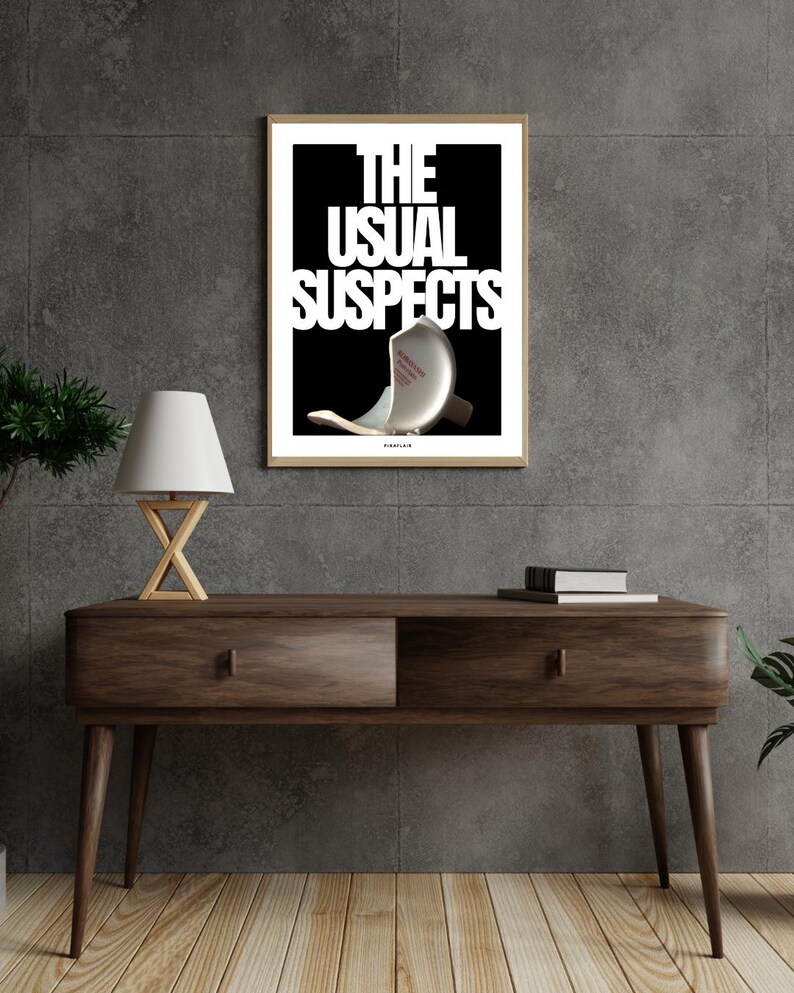 May include: A framed art print featuring the words "THE USUAL SUSPECTS" in white on a black backdrop. The image includes a broken object. The print is displayed on a dark wood console table with a lamp and books.