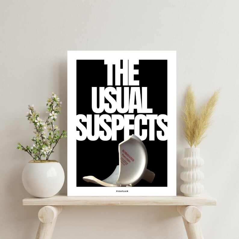 May include: A framed art print featuring the text "THE USUAL SUSPECTS" in white against a black backdrop. A broken ceramic bowl is in the foreground. The print is displayed on a wooden shelf, with decorative vases.