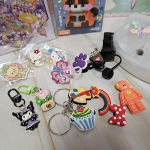 Puede incluir: Una colecci&oacute;n de llaveros y dijes coloridos con varios dise&ntilde;os. Incluye una magdalena, una paleta con un pincel, un arco&iacute;ris, una figura de superh&eacute;roe y personajes como Hello Kitty y Kuromi. Tambi&eacute;n incluye un tap&oacute;n de botella negro.
