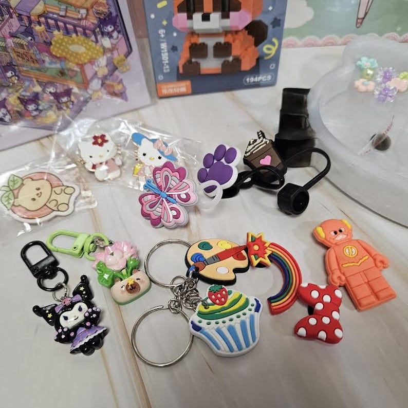 Puede incluir: Una colecci&oacute;n de llaveros y dijes coloridos con varios dise&ntilde;os. Incluye una magdalena, una paleta con un pincel, un arco&iacute;ris, una figura de superh&eacute;roe y personajes como Hello Kitty y Kuromi. Tambi&eacute;n incluye un tap&oacute;n de botella negro.