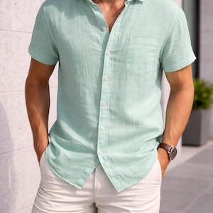 May include: A light green short-sleeved shirt with a chest pocket and white shorts. The shirt is buttoned and has a collar. A brown leather watch is visible on the wrist. The outfit is casual and suitable for warm weather.