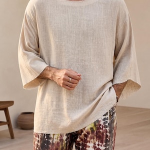 May include: A beige, loose-fitting top with a wide neckline and three-quarter sleeves. The top has a raw, unfinished edge. The model is wearing patterned trousers with brown, red, and white tie-dye designs. The outfit is casual and comfortable.