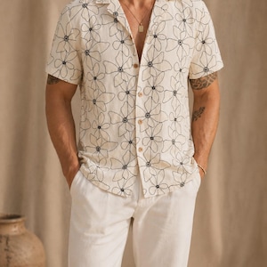 May include: Cream-colored short-sleeved shirt and white pants. The shirt features a black floral embroidery pattern and a button-down front. The outfit is a casual style, suitable for warm weather.