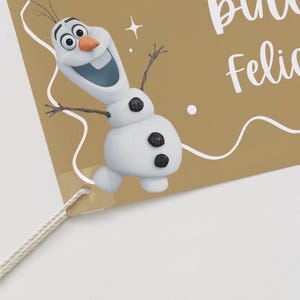 Puede incluir: Una etiqueta de color beige con una imagen de dibujos animados de Olaf el mu&ntilde;eco de nieve. La etiqueta tiene texto blanco que dice "Felicia". Olaf tiene una nariz de zanahoria, brazos de ramita y ojos de bot&oacute;n negros.