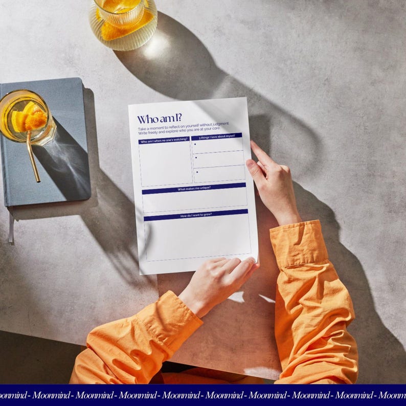 May include: A white sheet of paper with the text "Who am I?" in a dark blue font. The paper is held by a person wearing an orange long-sleeved shirt. A glass of orange-infused water and a blue notebook are also visible.