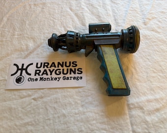 May include: A teal and yellow raygun with a detailed design, featuring a trigger and a scope. The raygun is placed on a white surface next to a sticker that reads "URANUS RAYGUNS One Monkey Garage."
