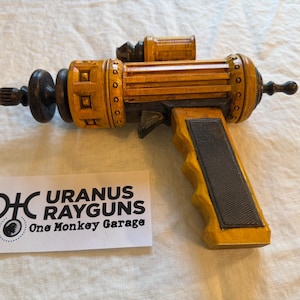 Puede incluir: Una pistola de rayos artesanal con un acabado amarillo y negro. La pistola presenta detalles intrincados, incluyendo un gatillo, una mira y varios acentos circulares y rectangulares. El texto "URANUS RAYGUNS" es visible en una etiqueta blanca.