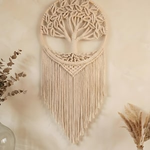 May include: A beige macrame wall hanging with a tree design. The tree is within a circular wooden frame, with knotted branches and leaves. Long, fringed strands hang below, creating a textured, bohemian aesthetic. The piece is designed for wall decor.