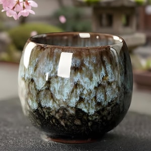 Puede incluir: Una taza de té de cerámica con una superficie brillante y texturizada. La taza presenta una mezcla de colores marrón, azul y negro, creando un efecto veteado único. La taza tiene una forma redondeada y una base pequeña.