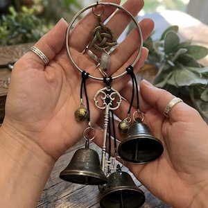 Könnte beinhalten: Ein handgefertigter Windspiel mit einem großen silbernen Reifen, einem keltischen Knotenanhänger, einem Schlüssel und drei dunklen Metallglocken. Kleine goldfarbene Glocken und ein Edelstein sind ebenfalls angebracht. Das Windspiel ist an schwarzen Kordeln aufgehängt.