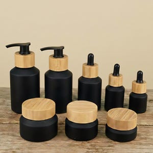 Peut inclure: Ensemble de flacons et pots en verre noir avec des couvercles en bambou naturel. L'ensemble comprend des flacons à pompe, des flacons compte-gouttes et de petits pots, adaptés aux soins de la peau ou aux huiles essentielles. Les contenants sont disposés sur une surface en bois.