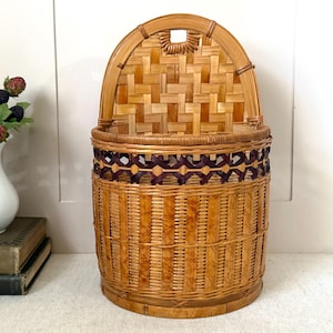May include: A vintage woven basket with a rounded top and a hinged lid. The basket is made of light brown and dark brown woven bamboo. The top section features a decorative woven pattern, and the body has vertical woven strips.