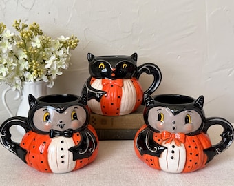 May include: Three ceramic mugs shaped like stylised creatures with black ears, orange and white polka-dot bodies, and black handles. Two mugs feature grey faces with bow ties, while the central mug has a black face with an orange bow tie.