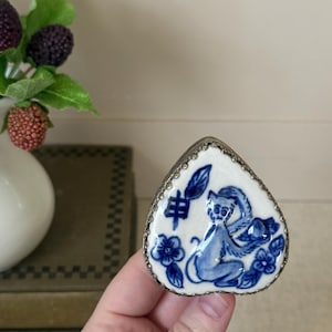 May include: Heart-shaped trinket box with a blue and white monkey design. The ceramic box has a silver-colored metal trim. The monkey is surrounded by blue floral accents and Chinese characters. The box is held in a hand.