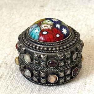 May include: A small, round, antique silver box with a colorful, mosaic-style lid. The box is adorned with gemstones and intricate metalwork, featuring a detailed pattern of butterflies and beads. The lid showcases a vibrant floral design.