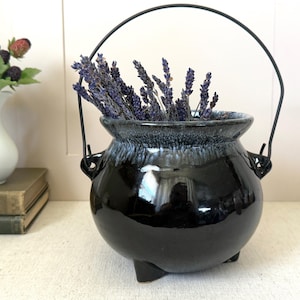 May include: A glossy black ceramic cauldron with a dark blue and white rim, holding dried lavender. The cauldron has a black metal handle and three small feet. A white vase with berries is in the background.