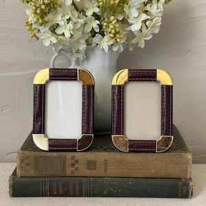 May include: Two rectangular picture frames featuring dark burgundy leather-like borders with visible stitching and polished gold-tone metal corner accents. The frames are positioned upright, side-by-side, displaying a blank white center area.