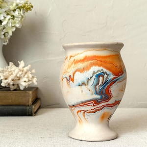 May include: A matte off-white ceramic vase with a flared rim and pedestal base, featuring a marbled pour paint design in vibrant shades of orange, blue, and black that creates swirling, fluid patterns across the exterior surface.