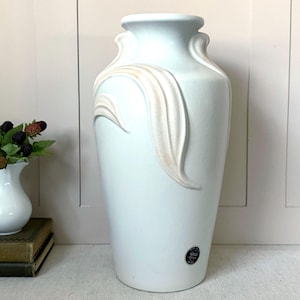 May include: A tall, matte white ceramic vase featuring a raised, flowing leaf-like relief design on its upper body. The vase has a wide, flared rim and a small, circular black and white sticker near the base with visible text.