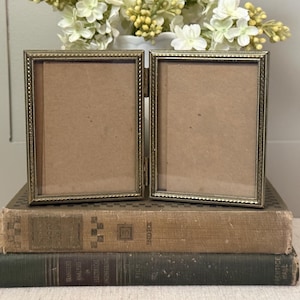May include: A hinged, antique-style, gold-toned metal picture frame with two rectangular openings. The frame is resting on two stacked, aged books. White and yellow flowers are in the background.